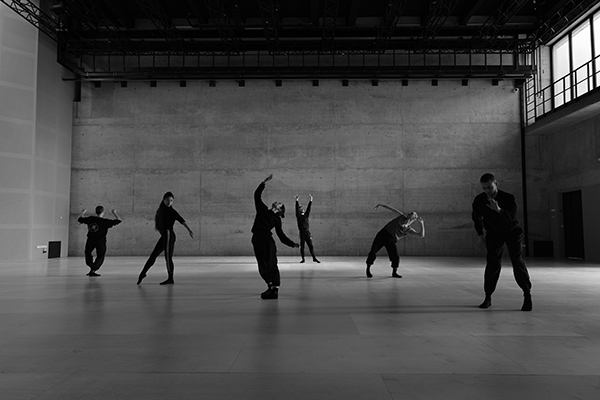 Czarno-białą fotografia przedstawia grupę tancerzy w trakcie zajęć, w odnowionej przestrzeni. Każdy z nich prezentuje inną figurę. Zdjęcie pochodzi z archiwum beneficjenta.