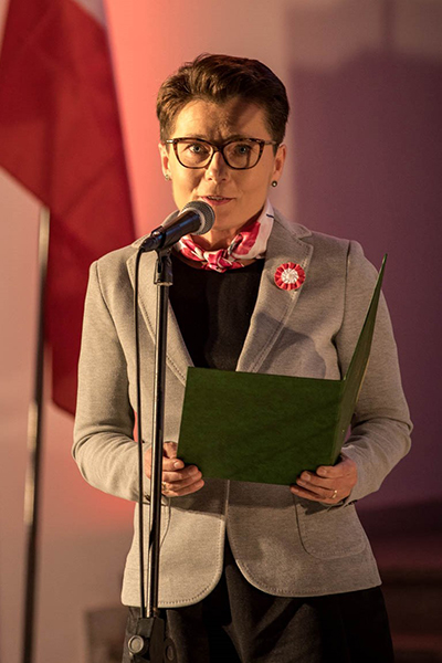 Fotografia przedstawia Danutę Potrawiak. Kobieta o krótkiej fryzurze, w okularach i jasnym żakiecie, trzyma w rękach folder z tekstem przemówienia. Przed nią ustawiony jest mikrofon. Zdjęcie pochodzi z archiwum prywatnego.