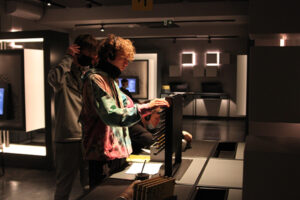 Fotografia przedstawia dwóch młodych chłopaków, którzy w interaktywnym muzeum rozwiązują zagadkę związaną z szyfrowaniem, na specjalnym urządzeniu. Autorem zdjęcia jest Dominik Wójcik.
